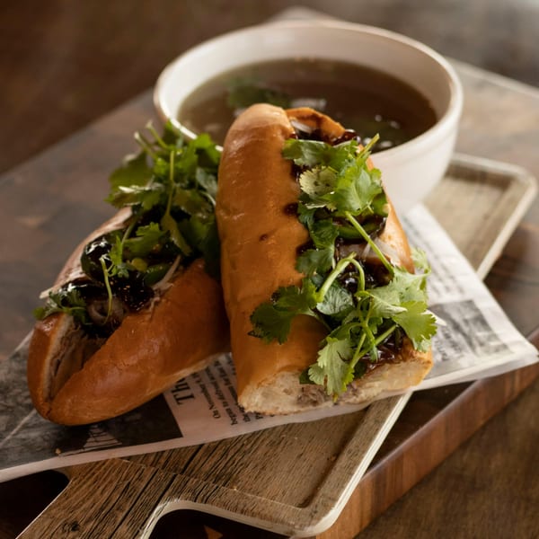 Two Vietnamese banh mi sandwiches with cilantro and sauce, served with a bowl of pho broth.