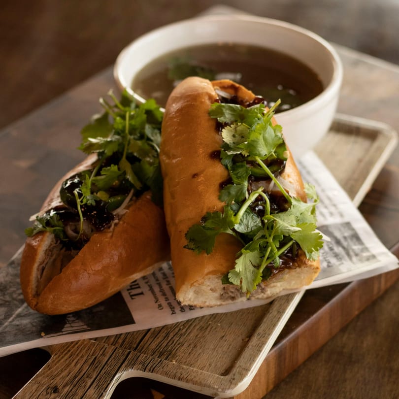 Two Vietnamese banh mi sandwiches with cilantro and sauce, served with a bowl of pho broth.