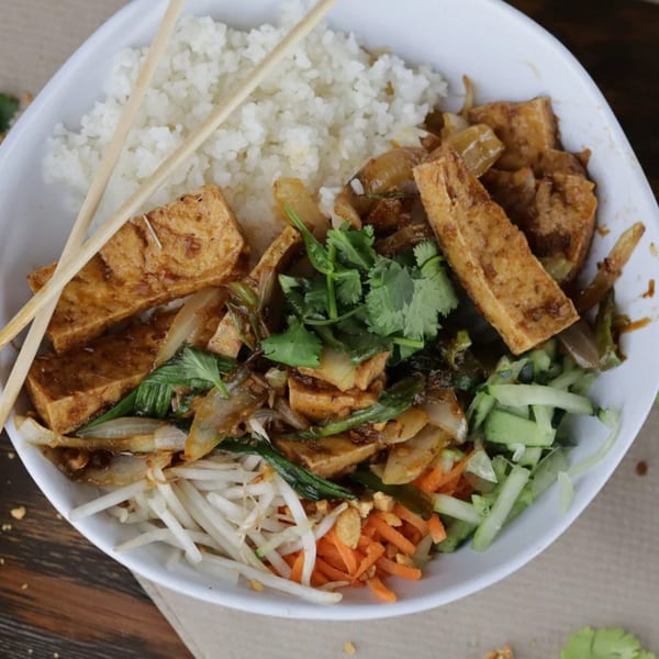 A white bowl of Vietnamese food with rice, tofu, vegetables, and chopsticks.