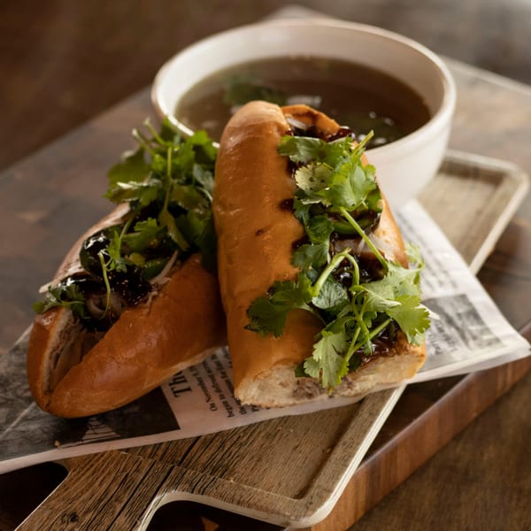 Two Vietnamese banh mi sandwiches with cilantro and dark sauce, served with a bowl of broth.
