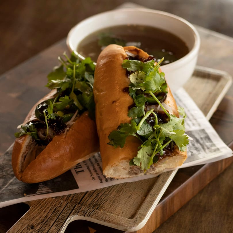 Two Vietnamese banh mi sandwiches with cilantro and dark sauce, served with a bowl of broth.