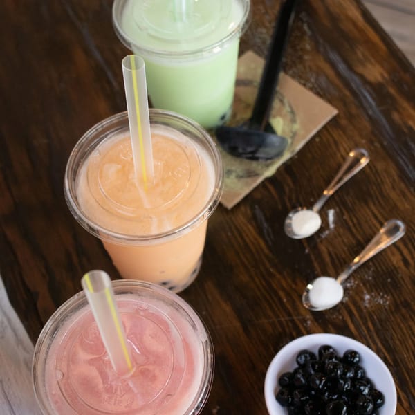 Three colorful bubble teas, a bowl of tapioca pearls, and spoons with sugar on a wooden table.