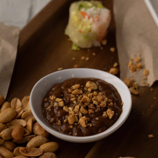 Peanut sauce with crushed peanuts, whole peanuts, and a spring roll on a wooden board.