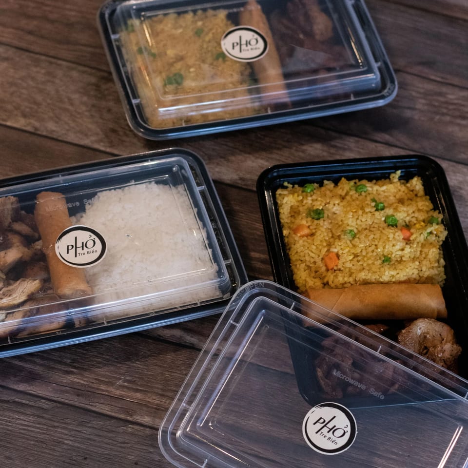 Three Pho Tre Bien takeout containers with Vietnamese food, including fried rice, white rice, and spring rolls.