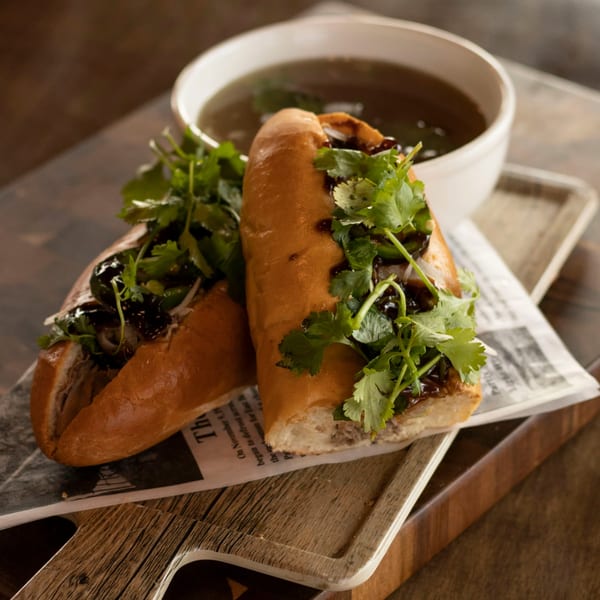 Two Vietnamese banh mi sandwiches with cilantro and dark sauce, served with a bowl of broth.