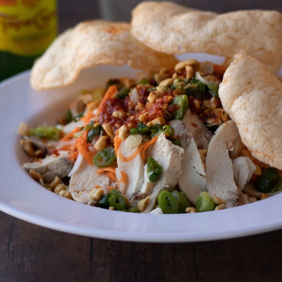Vietnamese chicken salad with peanuts, chili sauce, and shrimp crackers on a white plate.