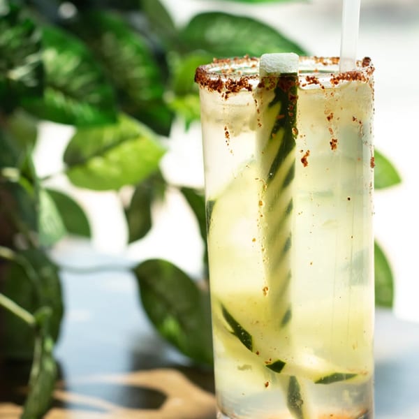 Tall glass of light yellow drink with cucumber slices, chili-lime rim, and a straw.