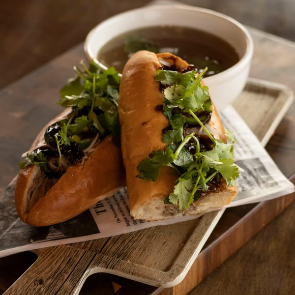 Two Vietnamese banh mi sandwiches with cilantro and sauce, served with a bowl of broth.
