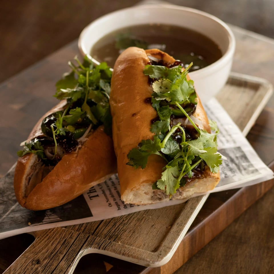 Two Vietnamese banh mi sandwiches with cilantro and sauce, served with a bowl of broth.