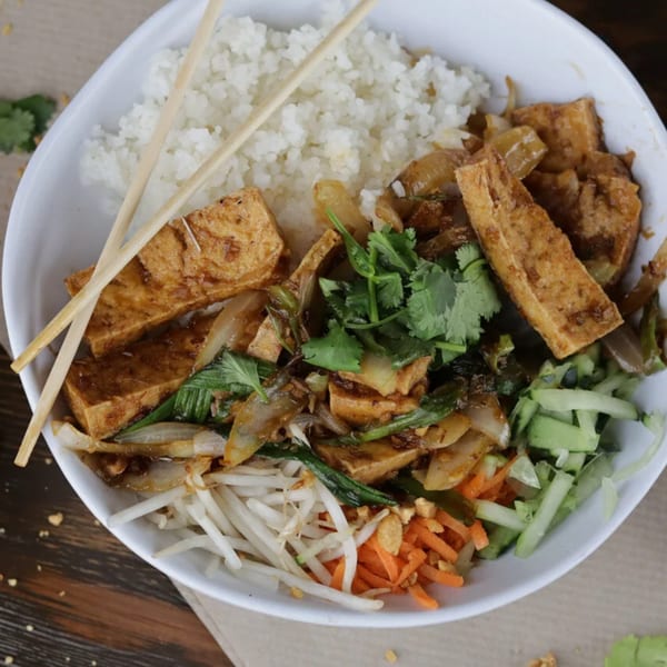 A white bowl of Vietnamese food with rice, tofu, vegetables, and chopsticks.