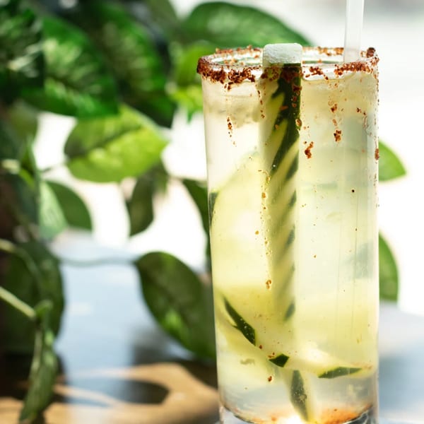 Tall glass of light yellow drink with cucumber, chili-lime rim, and a straw, against green foliage.
