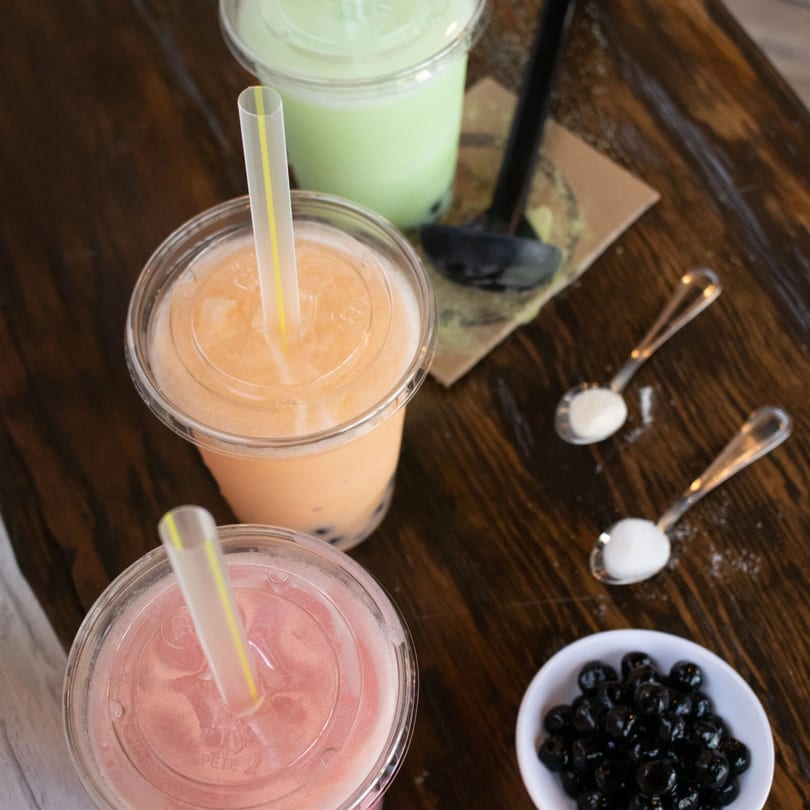 Three colorful bubble teas, tapioca pearls, and sugar on a wooden table.
