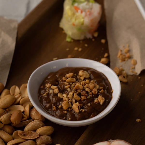 A white bowl of peanut sauce topped with crushed peanuts, next to whole peanuts and a spring roll.