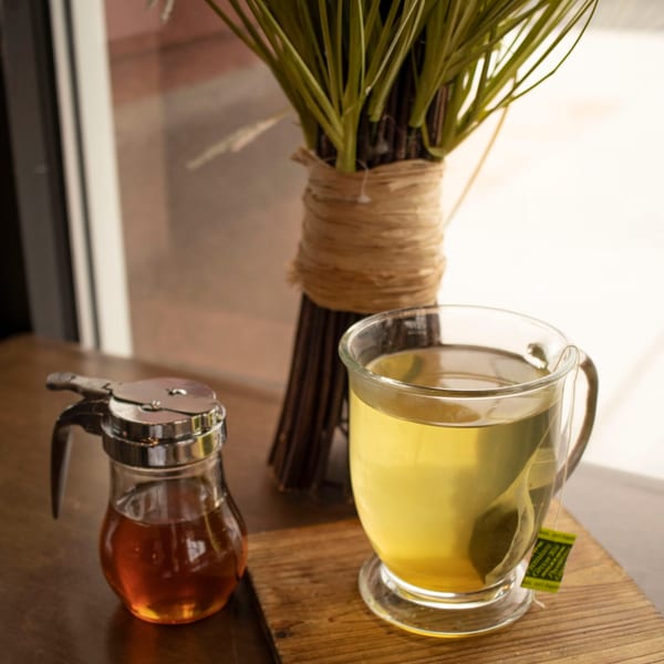 A glass of hot green tea with a tea bag and a honey dispenser on a wooden table.